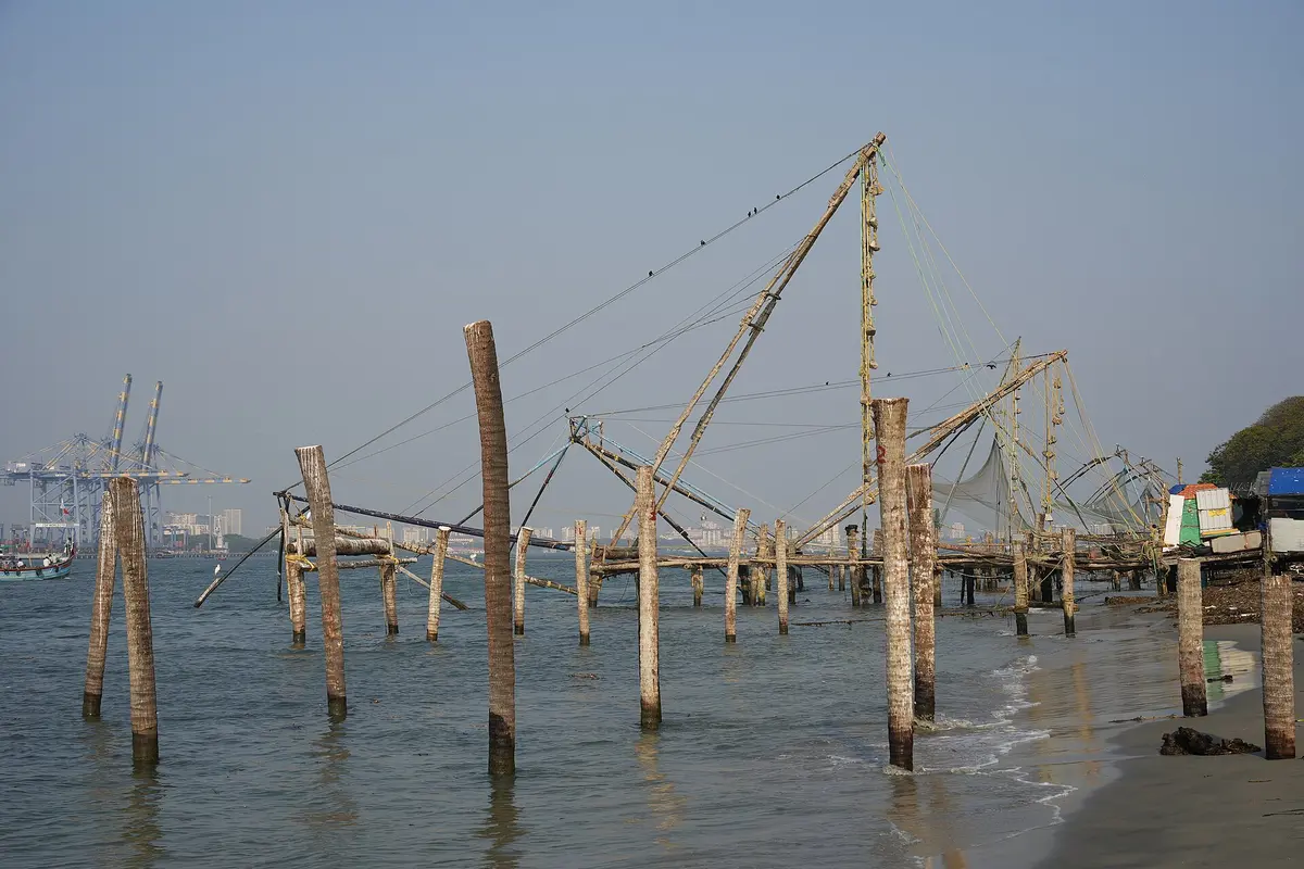 Chinese fishing nets on wooden poles at the Fort Kochi waterfront, with the sea in front and port cranes in the distance