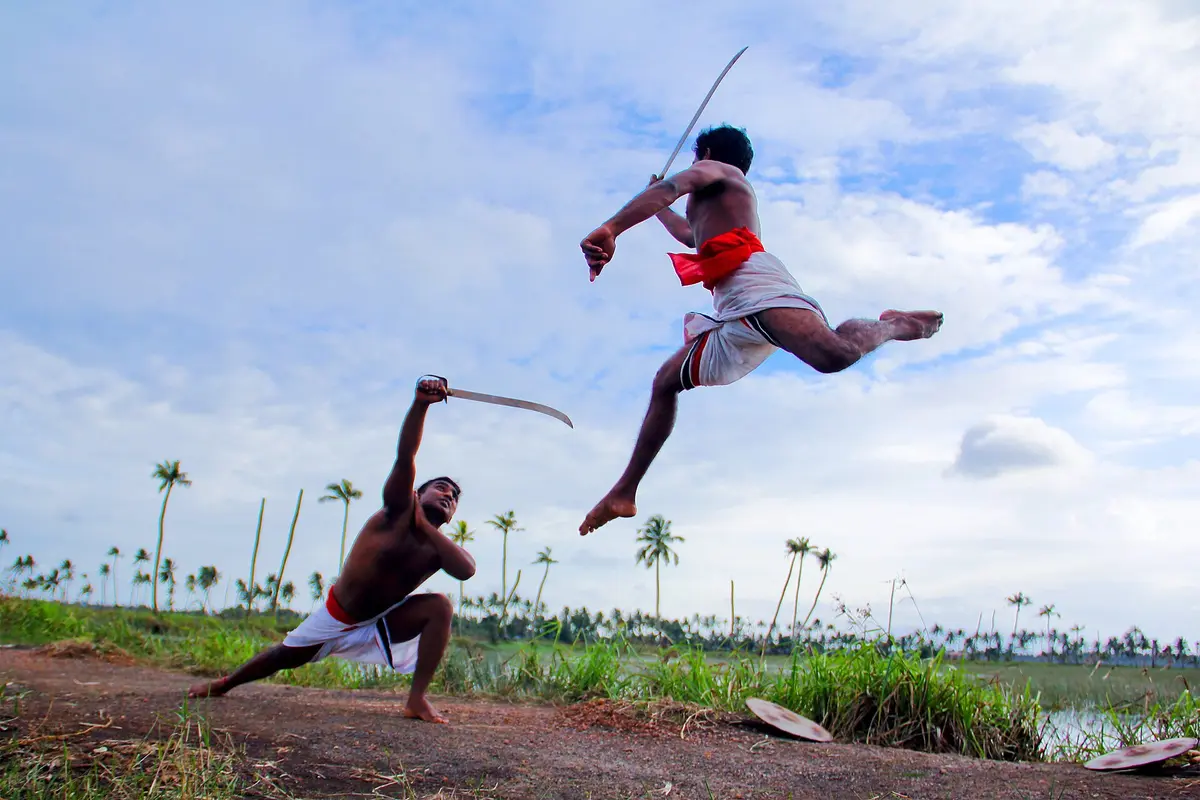 Two Kalaripayattu swordsmen in mock combat on open ground, one leaping in the air, with coconut palms behind