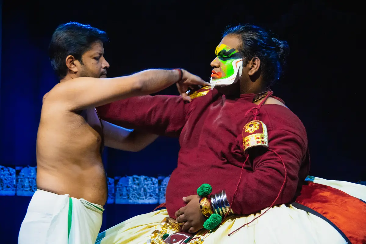 A Kathakali actor with green and white painted face being dressed on a dark stage by an assistant