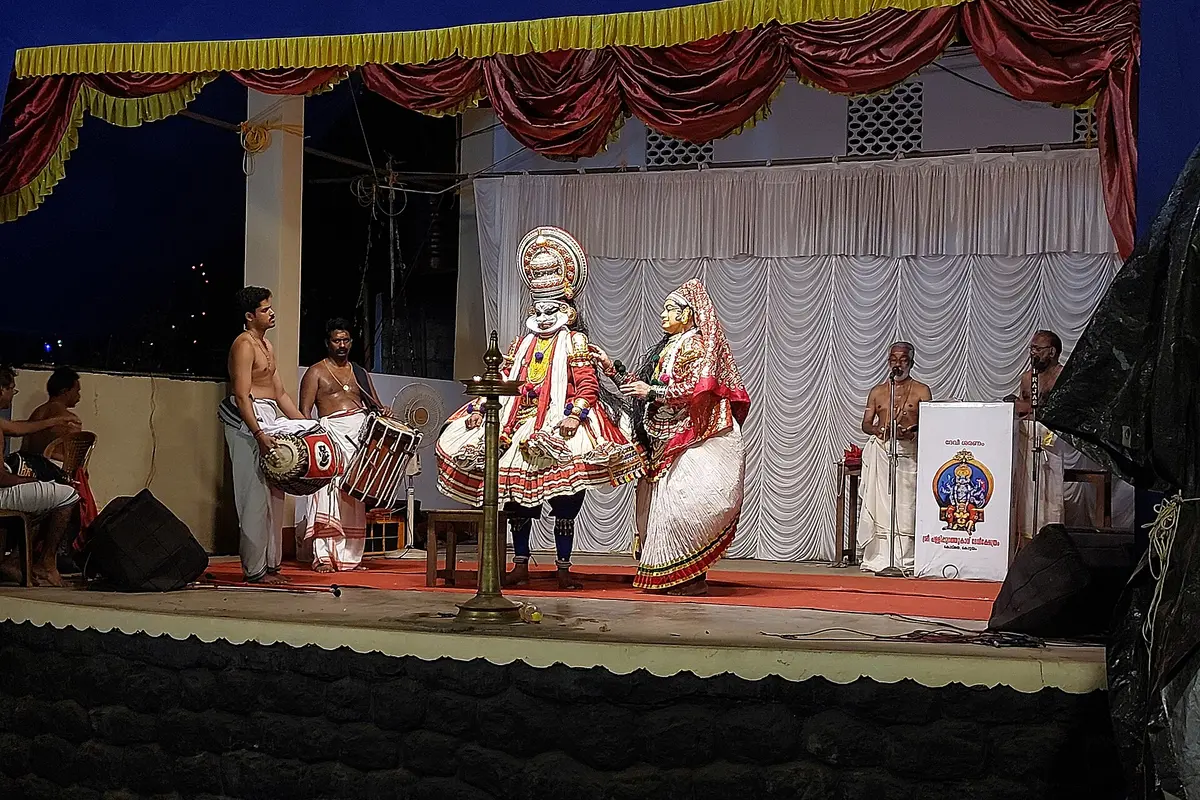 Two Kathakali actors in costume on a temple-festival stage around a brass oil lamp, with drummers on the left and singers on the right
