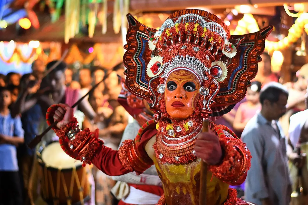 A Theyyam performer in a tall red and silver headdress with a painted orange face, drummers and a crowd behind